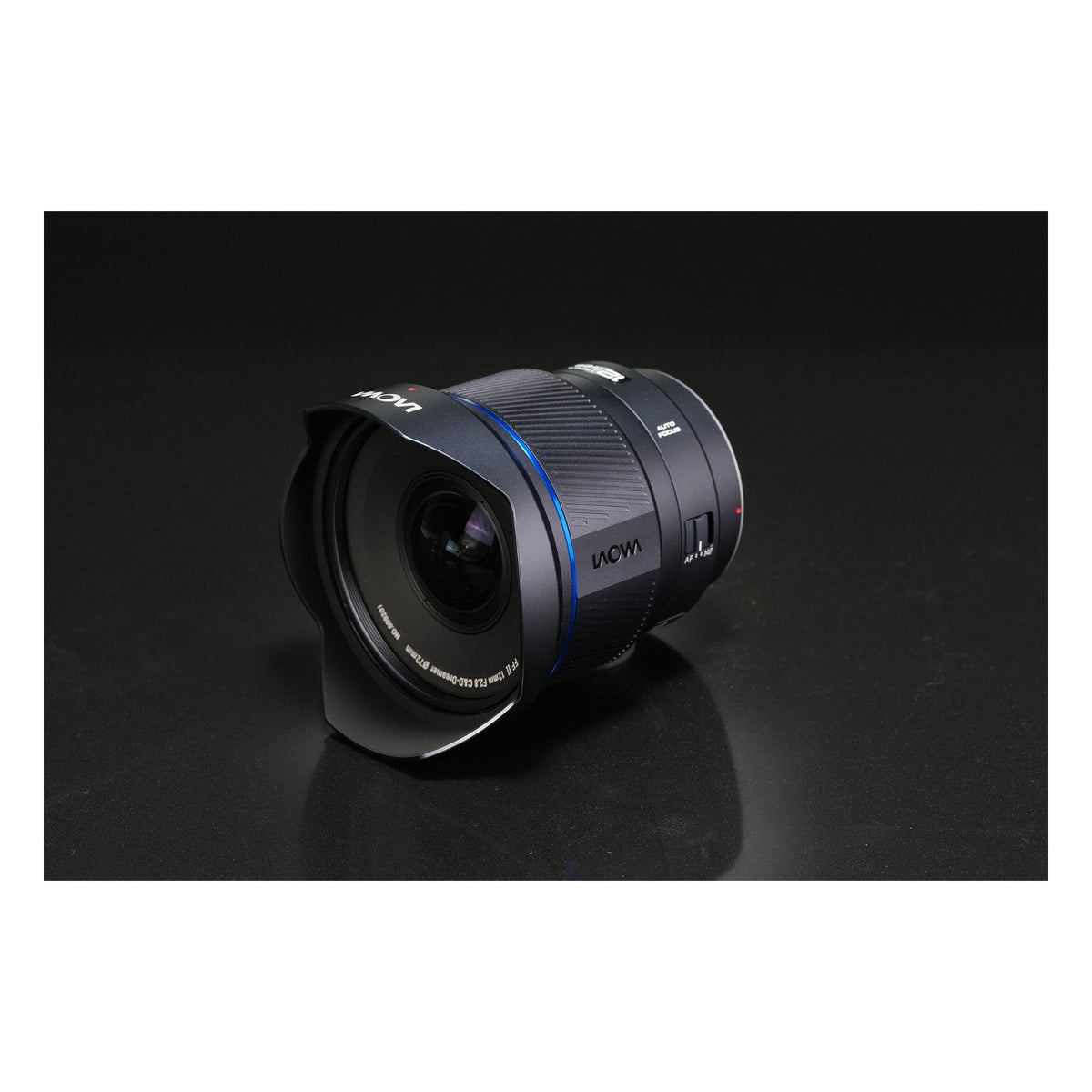 Laowa 12mm f/2.8 reclining at an angle on a dark grey surface with subtle reflections, showing side profile and optical elements from a relaxed perspective.