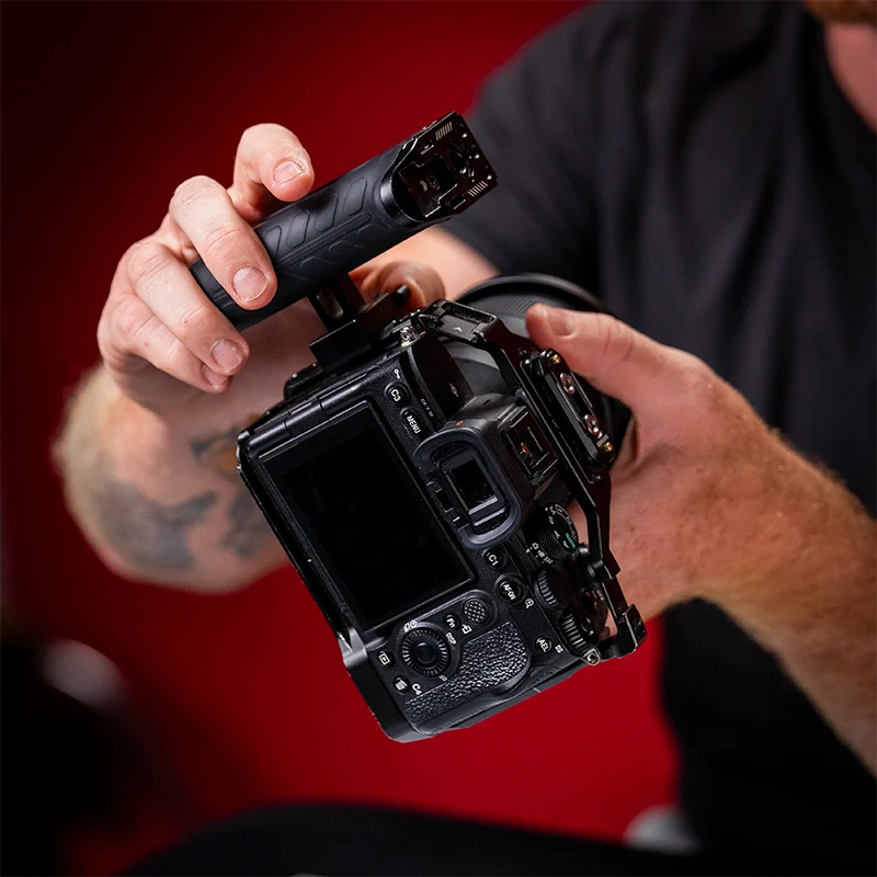 Person attaching a SmallRig side handle to a Sony A7 IV camera housed in a SmallRig cage, with a red studio background.