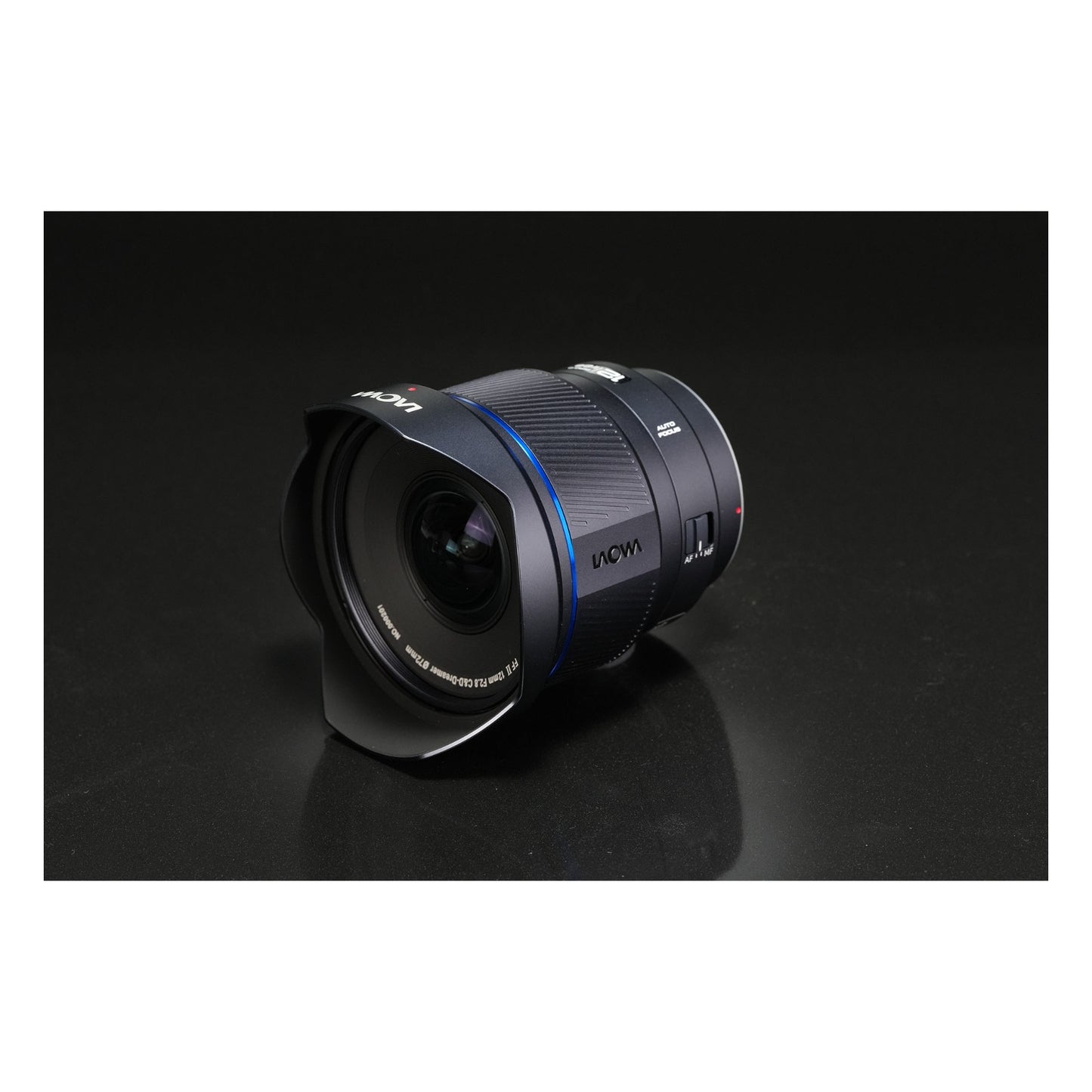 Laowa 12mm f/2.8 reclining at an angle on a dark grey surface with subtle reflections, showing side profile and optical elements from a relaxed perspective.