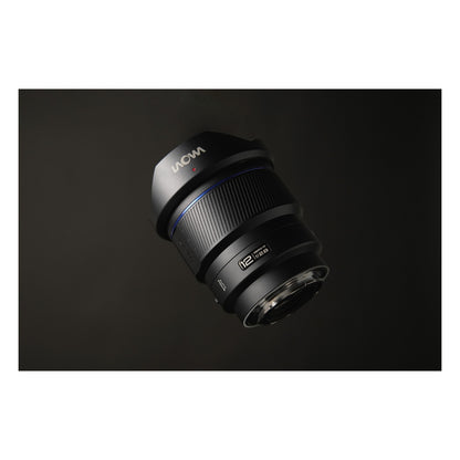 Laowa 12mm f/2.8 photographed from above against a dark gradient background, hovering position emphasizing the barrel geometry and hood form with even, subtle side-lighting.
