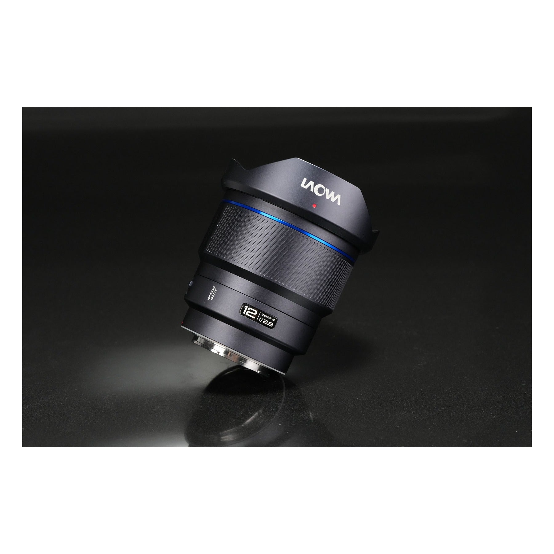 Laowa 12mm f/2.8 tilted upright on dark surface with hood deployed, captured with moody sidelighting that emphasizes lens texture and the blue accent ring.