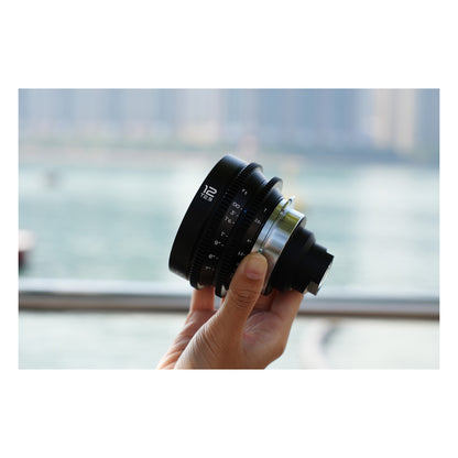 Laowa 12mm T2.9 Lite Zero-D lens held in palm outdoors with blurred water/cityscape background, showing compact scale and handheld ergonomics.